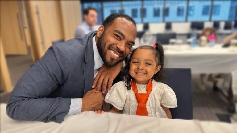 Justyn Turner in a suit, smiles while sitting next to a young girl wearing an oversized orange tie. They are seated at a table covered with white tablecloths in a brightly lit room with large windows.