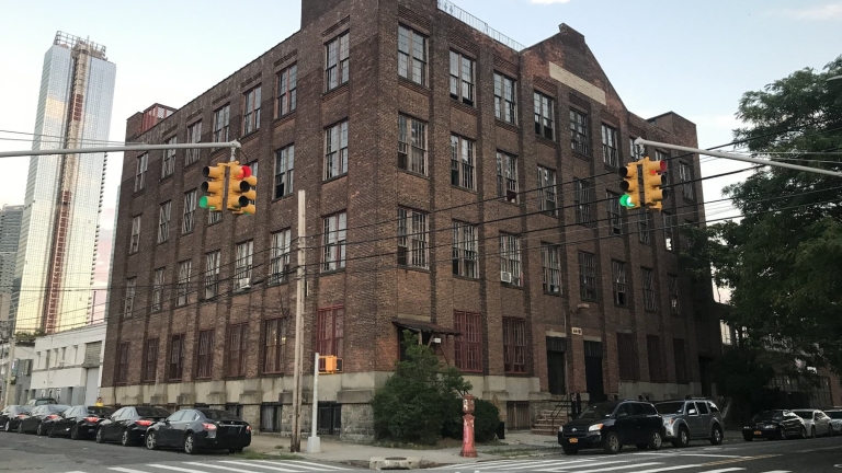 Four-story Metropolitan Building on Long Island City corner with traffic lights and parked cars. Tall modern skyscraper in the background. Cloudy sky.