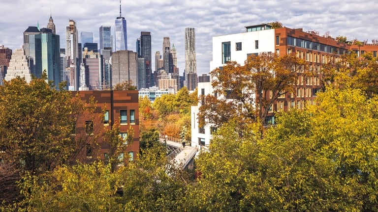 View of Squibb Park Bridge in Brooklyn with the New York City skyline, tall buildings and skyscrapers in the background, and trees and mid-rise residential buildings in the foreground on a partly cloudy day.