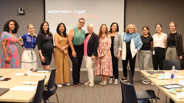 A group of eleven people stands in front of a presentation screen displaying "women.nyc NETWORK." They are smiling and dressed in casual to business-casual attire. A group of eleven people stands in front of a presentation screen displaying "women.nyc NETWORK." They are smiling and dressed in casual to business-casual attire.