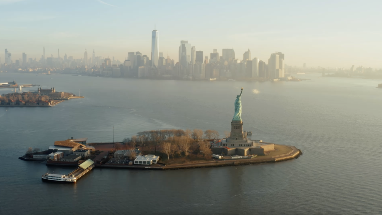 Aerial view of the Statue of Liberty on Liberty Island surrounded by water. In the background, the skyline of Manhattan, New York City, is visible with tall buildings under a clear sky. Aerial view of the Statue of Liberty on Liberty Island surrounded by water. In the background, the skyline of Manhattan, New York City, is visible with tall buildings under a clear sky.