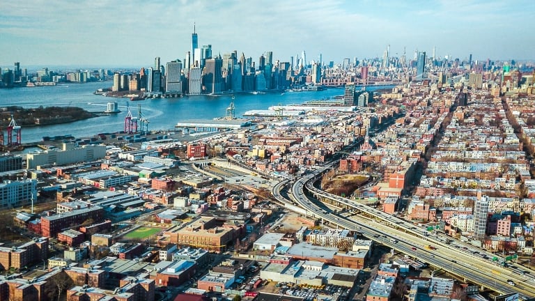 Aerial view of Red Hook Brooklyn featuring a mix of commercial, residential buildings, highways, and the New York Harbor with Manhattan skyscrapers in the background on a clear day.