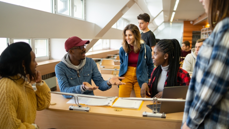 A group of students in a classroom engage in discussion, sitting and standing around desks with papers and a laptop.