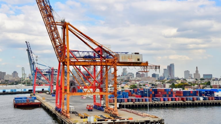 A large crane and shipping containers at Brooklyn Marine Terminal with NYC skyline in the background under a partly cloudy sky.