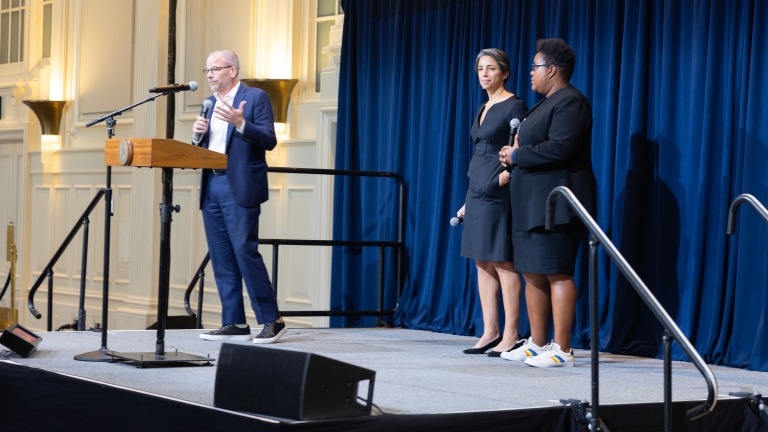 Three people standing on a stage at the NYC Climate Technology Showcase; Andrew Kimball is speaking at a podium, while the other two hold microphones.