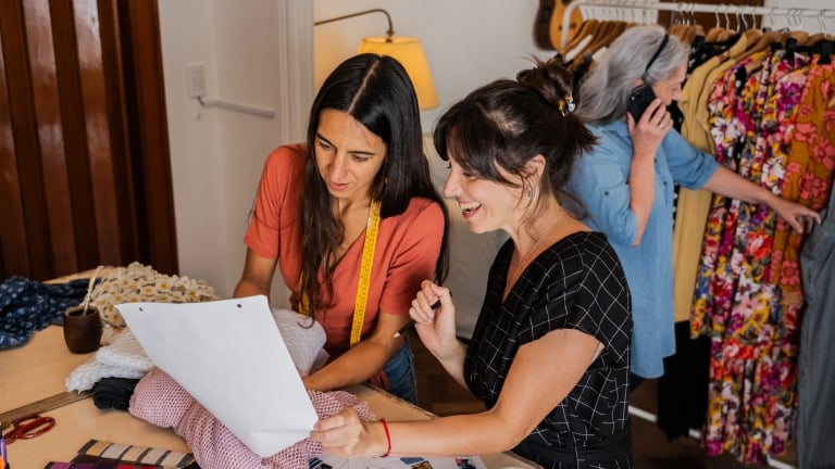 Two women fashion designers are looking at a sheet of paper and fabric samples in a sewing room. Another woman in the background is talking on the phone by a clothing rack.