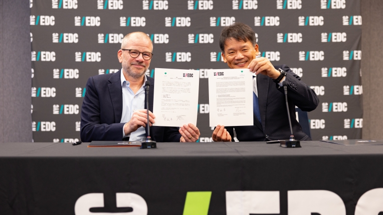 Two men seated at a table hold up signed MOU documents. A banner behind them displays the EDC logo repeatedly.