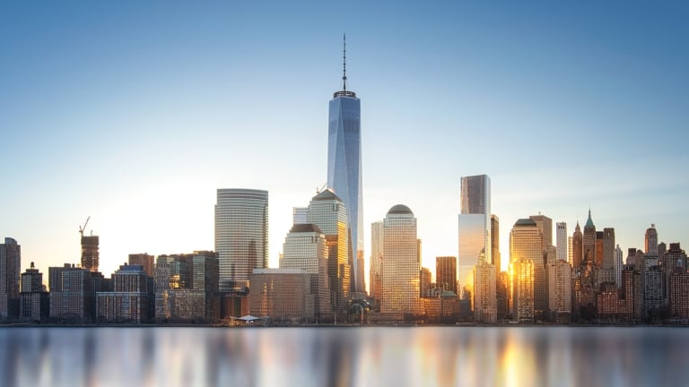 Skyline of New York City with modern high-rise buildings reflecting in calm water, under a clear blue sky at sunset.