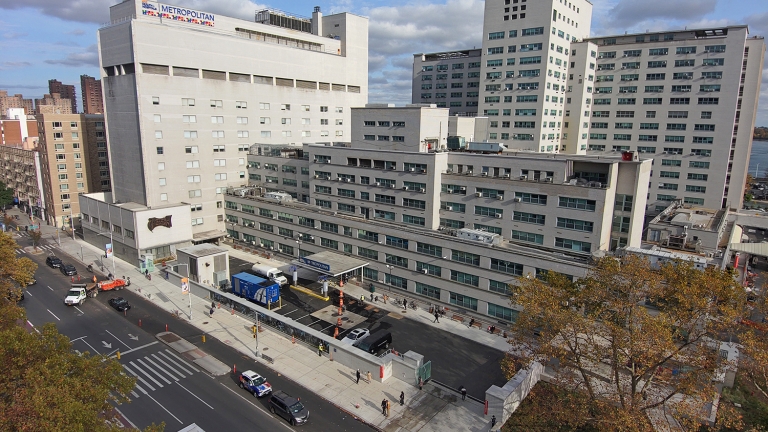 Aerial view of the large Metropolitan Hospital complex with multiple buildings and a busy street in the foreground. Trees with autumn foliage line the area.