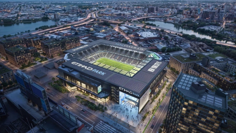 Aerial view of a football stadium labeled &quot;Etihad Park&quot; surrounded by urban landscape, with roads and a river in the background.