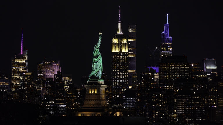 Night view of the New York City skyline featuring the illuminated Statue of Liberty in the foreground and skyscrapers in the background.