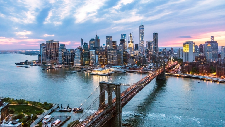 Aerial view of the Brooklyn Bridge leading to Manhattan, New York City, at dusk. The skyline is illuminated, reflecting on the water, with the horizon displaying a colorful sunset behind scattered clouds.