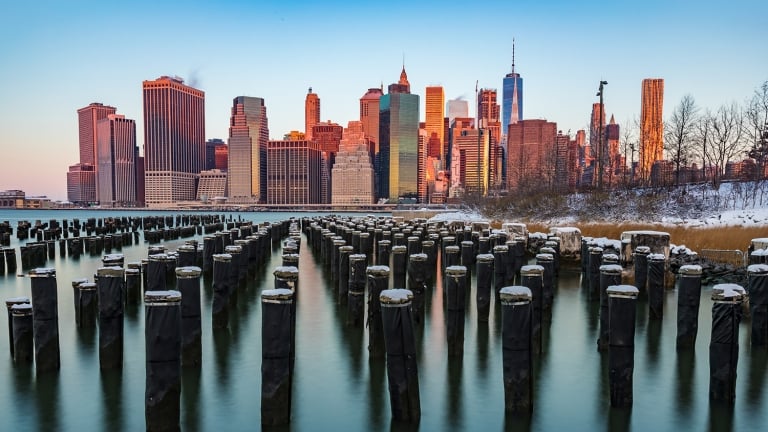 Manhattan skyline at sunrise viewed from the Brooklyn Bridge Park, with wooden pilings covered in snow in the foreground and a clear sky above.