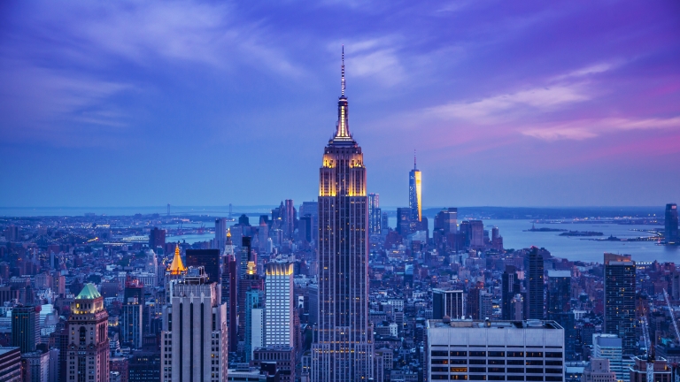 A scenic view of New York City skyline at dusk, featuring an illuminated Empire State Building in the center. The sky is a blend of purple and blue hues, with surrounding buildings and a body of water visible in the distance.