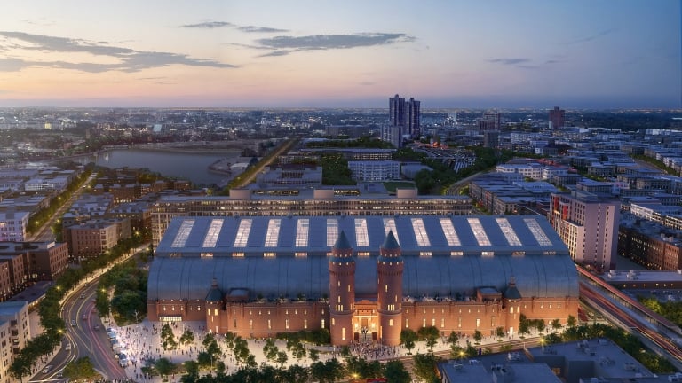 Aerial view of a large, illuminated Kingsbridge Armory building with two towers, surrounded by cityscape at dusk. The building features a glass-domed roof and is set against a cloudy sky with a pinkish hue. Streets and trees are visible around the structure.