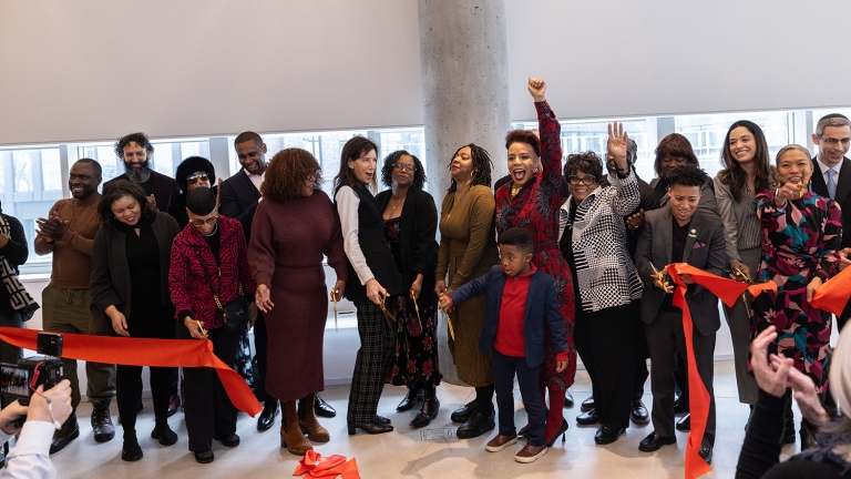 A diverse group of people celebrates at a ribbon-cutting ceremony inside a building. They hold a long red ribbon, and several individuals have their arms raised in excitement. Photographers are capturing the moment.