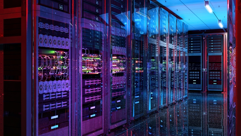 A modern server room with multiple server racks on either side. The racks are filled with cables and illuminated by colorful LED lights, casting a vibrant glow on the reflective floor. The ceiling has bright overhead lights.