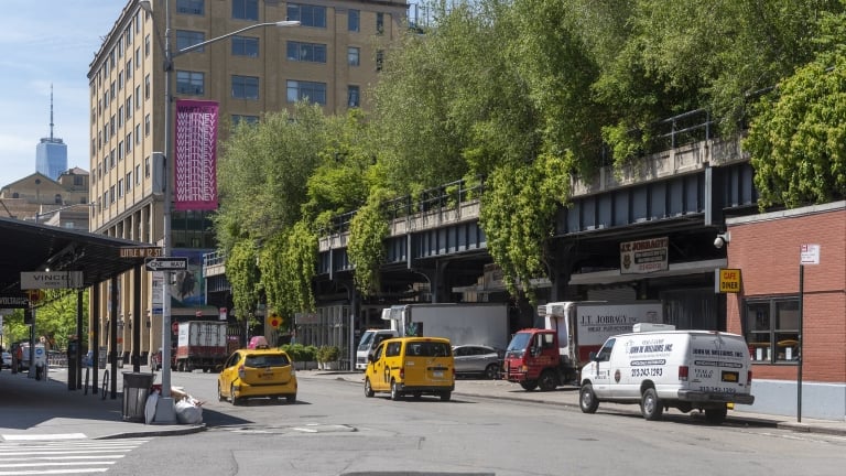 Street scene with a view of the High Line in New York City. The old railway track is lined with greenery and bordered by buildings. Yellow taxis, a van, and a café with a red awning are visible. The One World Trade Center is in the background. Street scene with a view of the High Line in New York City. The old railway track is lined with greenery and bordered by buildings. Yellow taxis, a van, and a café with a red awning are visible. The One World Trade Center is in the background.