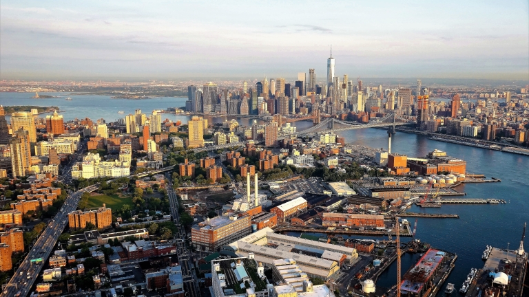 Aerial view of Manhattan, New York City, showcasing skyscrapers, the Hudson River, and surrounding boroughs. The One World Trade Center is prominently visible, with sunlit buildings and bustling waterways in the foreground.