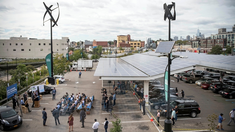 A parking lot features solar panels as a canopy. People are gathered for an event beneath the panels. Wind turbines are visible above, and there are banners and chairs set up. The background shows an urban landscape with buildings. A parking lot features solar panels as a canopy. People are gathered for an event beneath the panels. Wind turbines are visible above, and there are banners and chairs set up. The background shows an urban landscape with buildings.