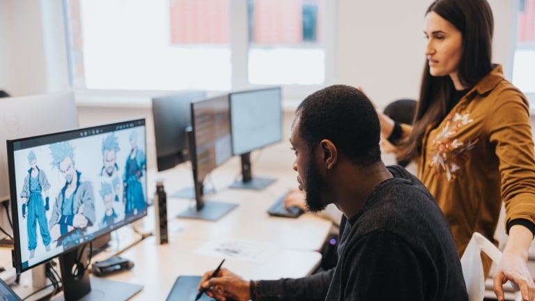 A man uses a drawing tablet and stylus while working on a character design on a computer screen in an office. A woman stands beside him, looking at the screen. Other monitors and desks are visible in the background.