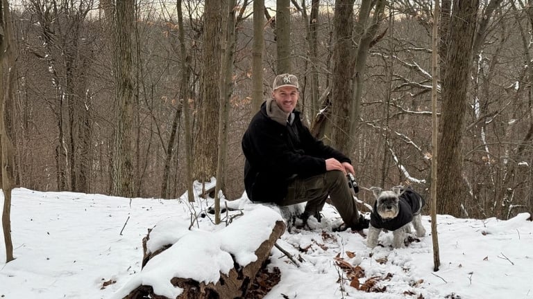 Matthew Furlong dressed in a black jacket and cap sits on a log in a snowy forest, holding a camera. Beside him is a small dog wearing a dark coat. Bare trees and a snowy landscape are in the background.