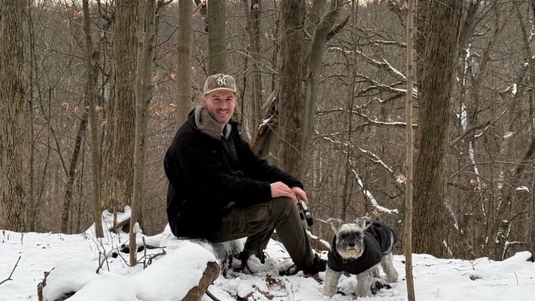 Matthew Furlong dressed in a black jacket and cap sits on a log in a snowy forest, holding a camera. Beside him is a small dog wearing a dark coat. Bare trees and a snowy landscape are in the background.