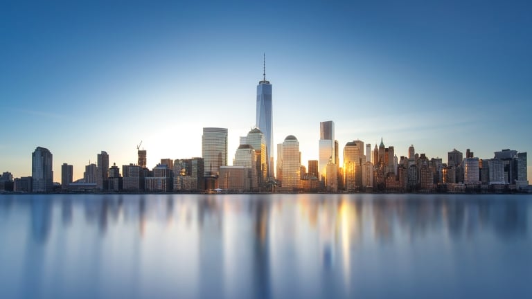 A panoramic view of a city skyline at sunset, featuring tall skyscrapers with the sun setting behind them, casting reflections on the calm water in the foreground. The sky is clear with a gradient from blue to warm tones near the horizon.