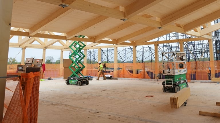 A construction site with a wooden roof structure. Workers are present, operating a green scissor lift and a smaller lift. Orange safety netting surrounds the area, and scaffolding is visible in the background. Building materials lie scattered on the floor.