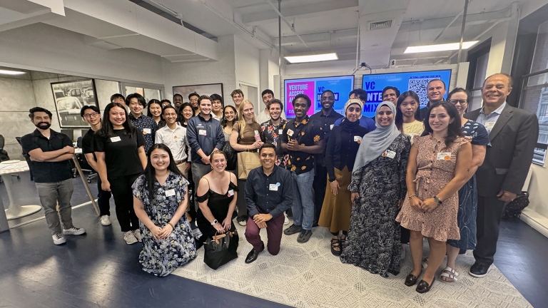 A diverse group of people, some standing and some kneeling, smile for a group photo in an indoor setting. They are at an event for the NYC Venture Capital Internship Program, with name tags visible on their clothing. A screen in the background adds context to the meeting.