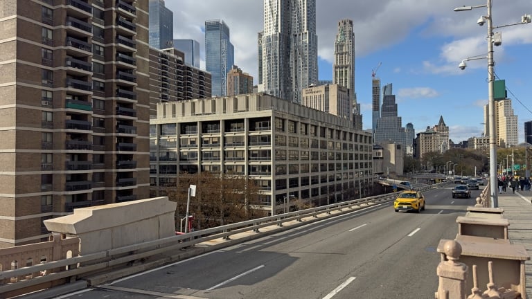 Urban scene at 100 Gold Street with a yellow taxi driving on a bridge in Manhattan. Tall skyscrapers are in the background beneath a partly cloudy sky. The setting appears busy and modern with multiple lanes on the road.