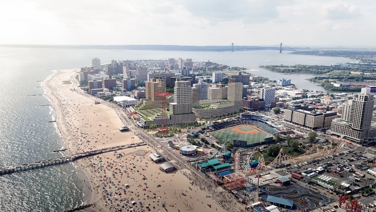 Aerial view of a Coney Island featuring a long sandy beach crowded with people, an amusement park with rides, a baseball stadium, and numerous buildings. The coastline curves with a distant bridge visible on the horizon.