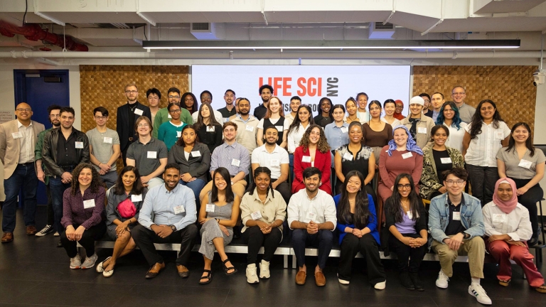 A diverse group of men and women smiling and posing for a group photo in a conference room. They are standing or sitting in front of a screen displaying Life Sci NYC. There are approximately 50 people in the image.