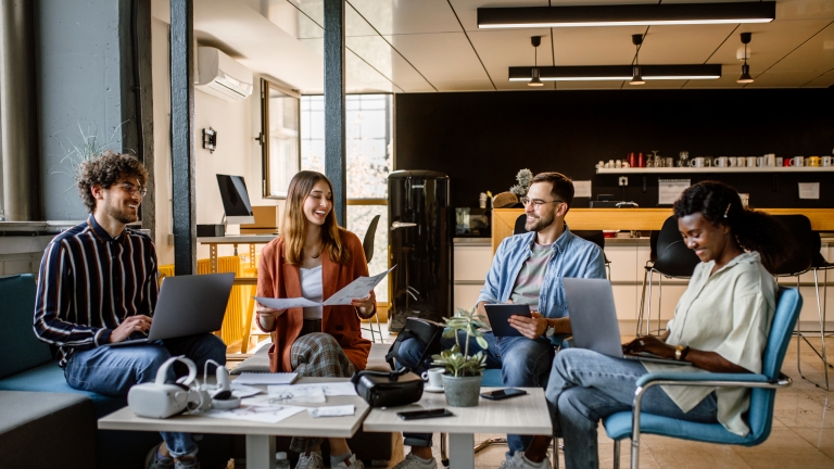 Four people sitting in a modern office, engaged in a casual meeting. They are using laptops and holding papers. The setting includes a coffee table, plants, and a kitchen area in the background, creating a relaxed atmosphere.