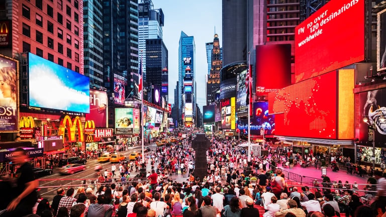 A bustling Times Square in New York City, filled with crowds and vibrant billboards. Bright lights from signs and advertisements illuminate the area. Yellow taxis are visible, and the skyline features tall buildings under a clear sky.