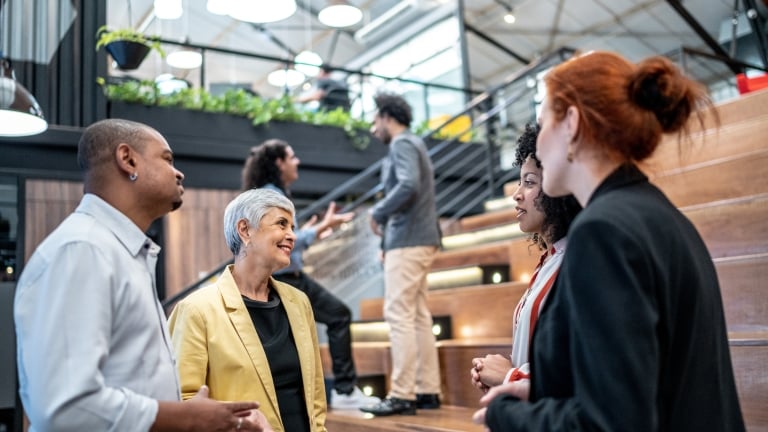 A group of diverse people engage in conversation on wooden bleachers in a modern office space. Green plants adorn the upper levels. Some people are standing while others sit or ascend the staircase in the background.