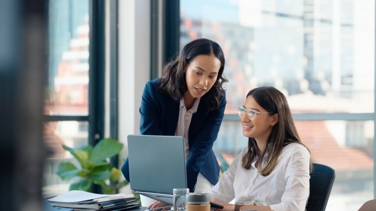 Two women in an office setting. One is standing and pointing at a laptop screen, while the other is seated, smiling and looking at the screen. There are notebooks and a coffee cup on the table. Large windows are in the background.