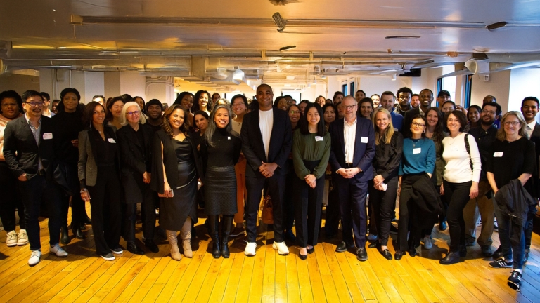 A diverse group of people stands closely together in a well-lit indoor space. They are smiling and dressed in business casual attire. The room has wooden floors and large windows in the background.