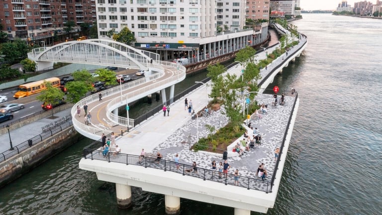 Aerial view of the East Midtown Greenway with a winding pedestrian bridge over the East River. People relax on benches surrounded by greenery. Adjacent is a city street with traffic, and tall buildings. A distant bridge is visible in the background.