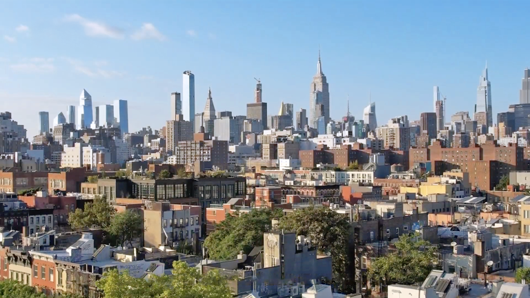 A cityscape view featuring a mix of modern skyscrapers and older buildings under a clear blue sky. Notable tall structures are visible in the distance, with greenery scattered among the foreground buildings.