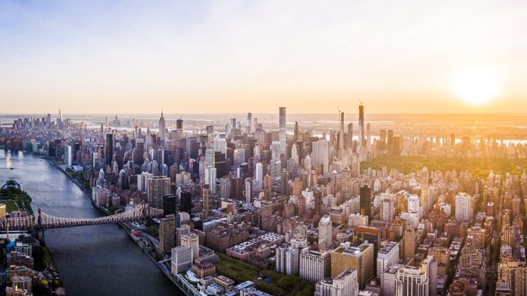 Aerial view of New York City skyline during sunset, featuring numerous skyscrapers and buildings. A wide river runs alongside the city, crossed by a prominent bridge. The sky is clear with the sun setting on the horizon.