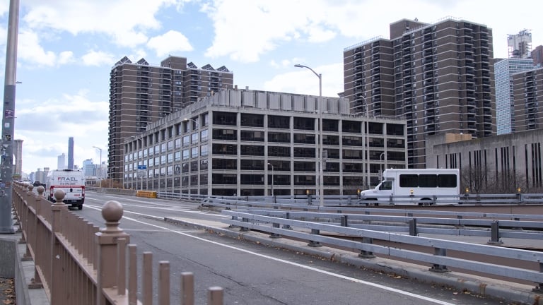 Urban scene showing a wide road with metal barriers on a bridge. A white van and a white truck are visible. Tall residential buildings with many balconies are in the background. The sky is partly cloudy.