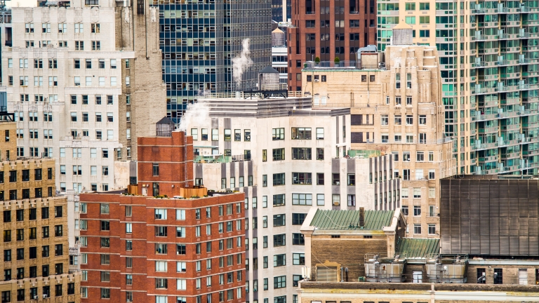 A close-up view of several tall buildings in a city, featuring a mix of brick, concrete, and glass exteriors with windows, rooftops, and some visible steam rising from a rooftop vent.