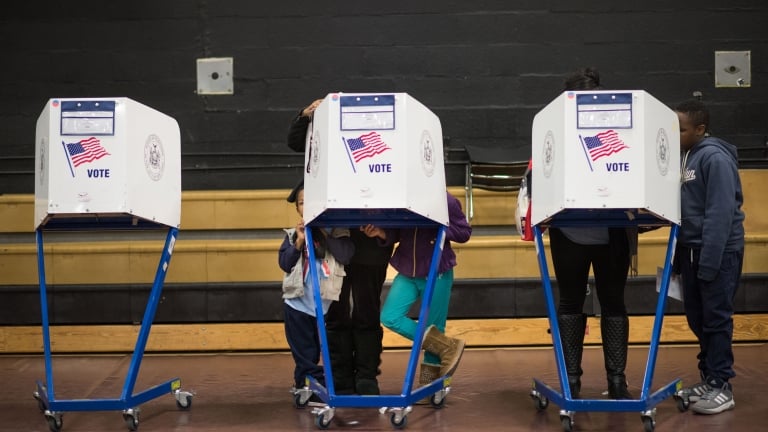 Three voting booths with “VOTE” and an American flag are occupied by people casting ballots in a gymnasium. Some children are visible, including one peeking from under a booth. Wooden bleachers are in the background.
