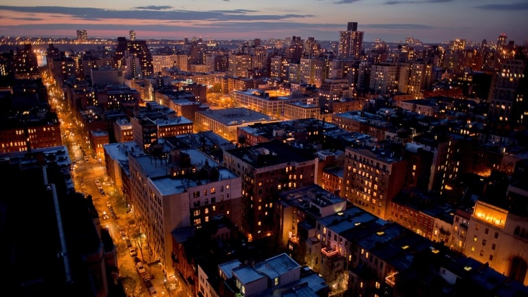 Aerial view of a city at dusk with illuminated buildings and streets, glowing windows, and a darkening sky with clouds on the horizon.