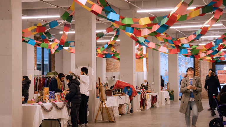 A spacious indoor market with colorful paper garlands hanging from the ceiling. Several people browse tables displaying various items like clothes, art, and crafts. A woman walks past with a stroller on the right.