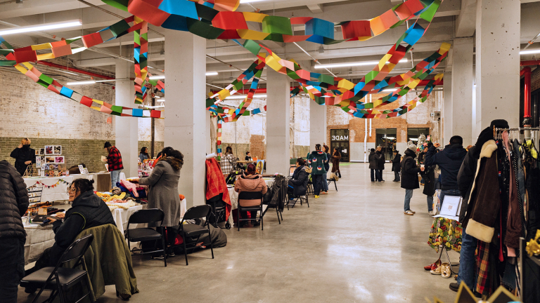 A spacious indoor market with people browsing vendor tables. Colorful paper garlands hang from the ceiling, and clothing racks and crafts are on display. The atmosphere is lively and festive.