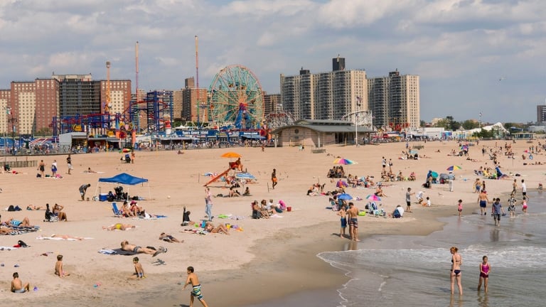 People relax on a sandy beach near the ocean, with colorful umbrellas, children playing in the water, and a cityscape with tall buildings, a Ferris wheel, and amusement park rides in the background. People relax on a sandy beach near the ocean, with colorful umbrellas, children playing in the water, and a cityscape with tall buildings, a Ferris wheel, and amusement park rides in the background.