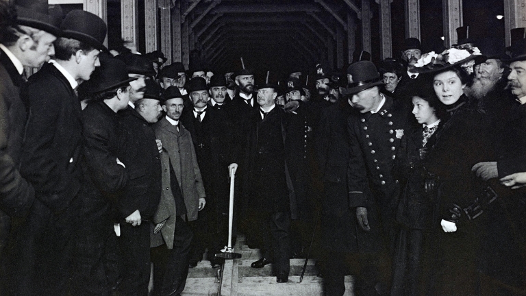 A black-and-white photo shows a crowd of men and women, some in hats and uniforms, gathered around a subway track inside a tunnel during a ceremonial event. Mayor Seth Low (center) hammers in spike for IRT subway, ca. 1902-04.