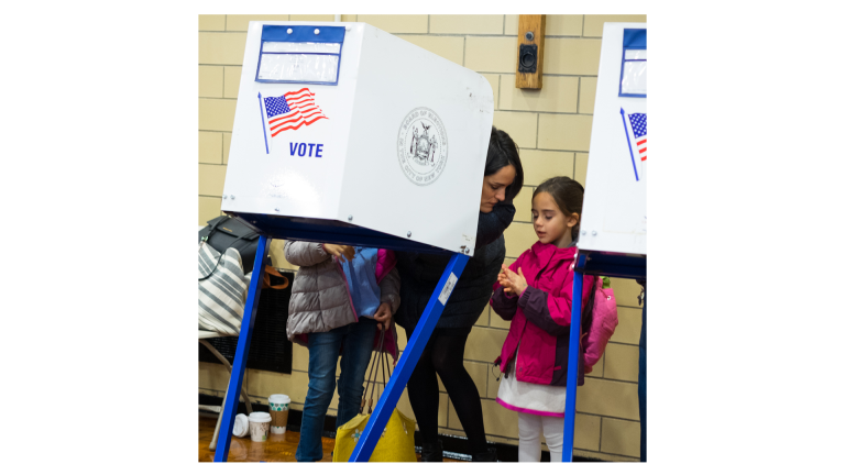 A woman and two children stand at a voting booth in a polling place. The woman appears to be voting while the children, both wearing jackets and backpacks, stand beside her. The booth displays the word VOTE and an American flag.